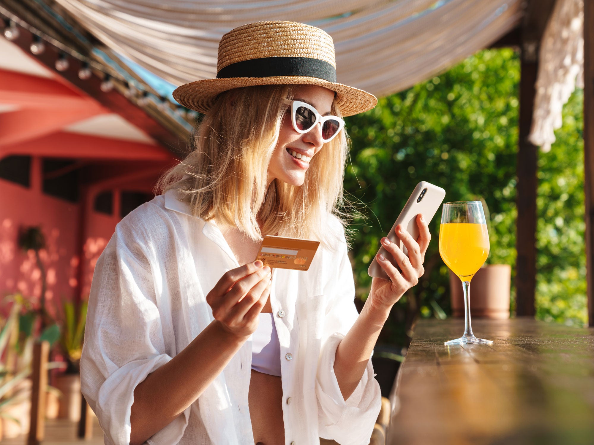 Eine Frau sitzt mit einem Getränk im Urlaub und hält eine KReditkarte in der einen und ihr Smartphone in der anderen Hand.