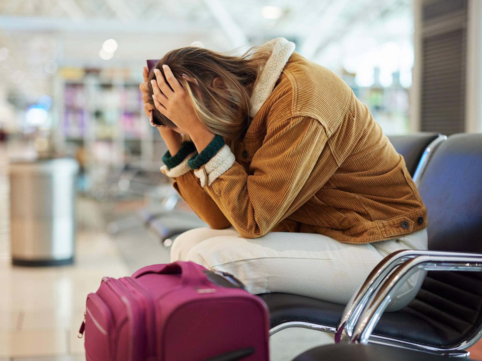 Frau sitzt mit gesenktem Kopf am Flughafen neben ihrem Koffer.