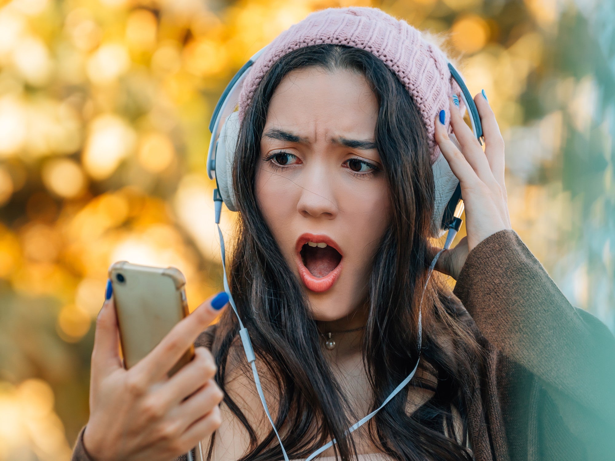 Frau mit Köpfhörern und Mütze schaut erschrocken auf ihr Handy-Display.