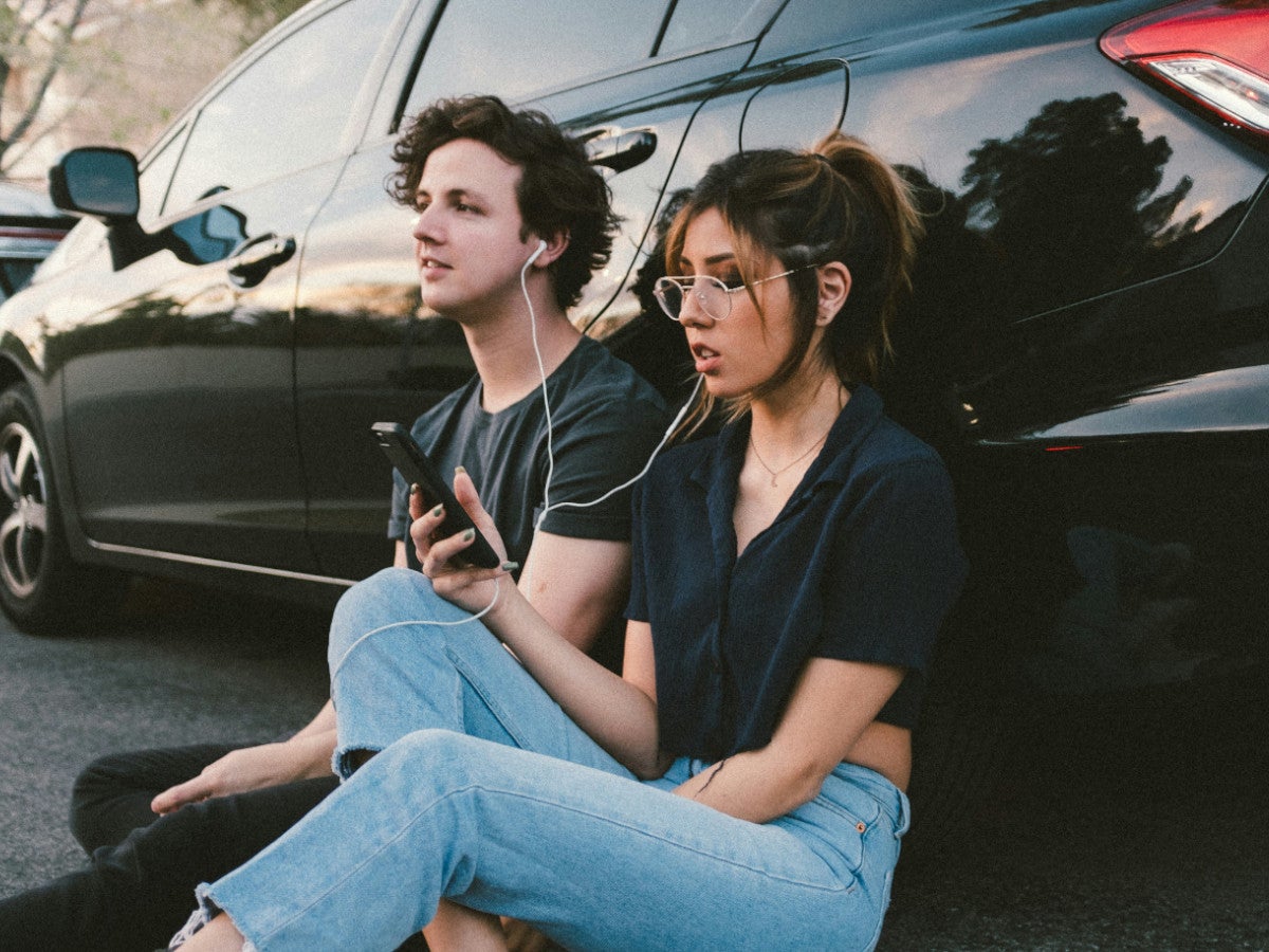 Ein Mann und eine Frau sitzen vor einem Auto und hören über Kopfhörer Musik.