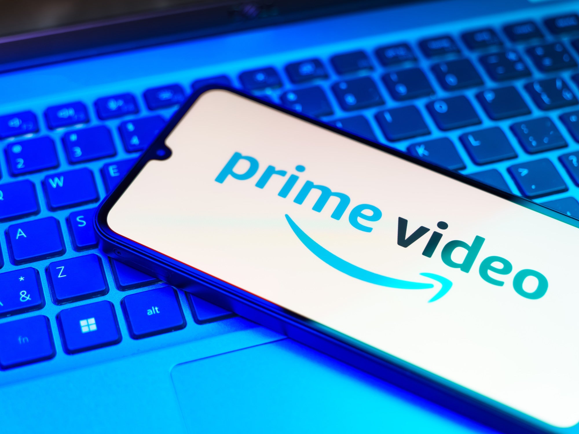 Logo von Prime Video auf einem Smartphone-Display das vor blauem Hintergrund auf einer Laptop-Tastatur liegt.