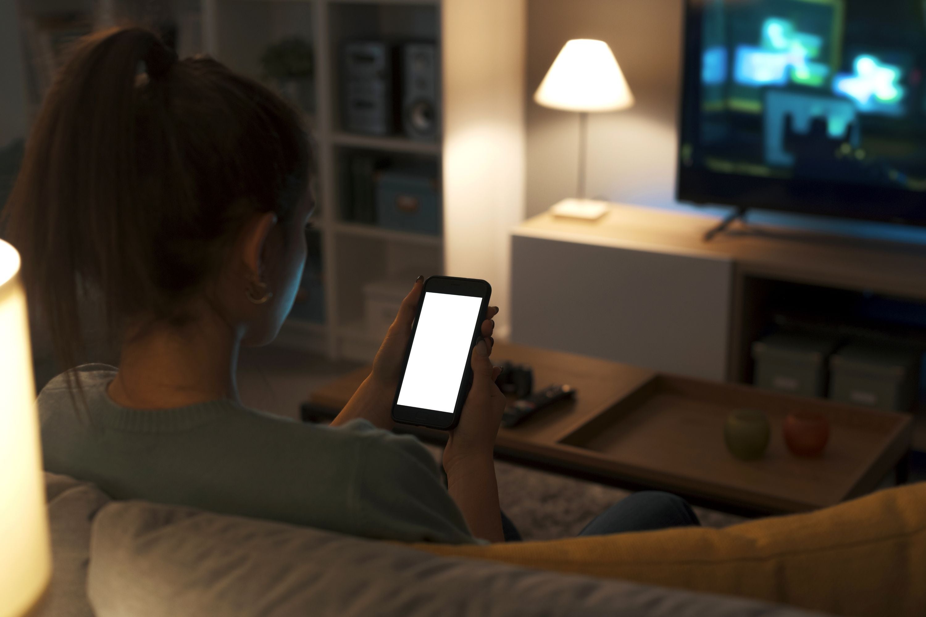 Ein Teenager sitzt auf einer Couch, hält ein Handy in der Hand. Im Hintergrund ist ein TV zu sehen.