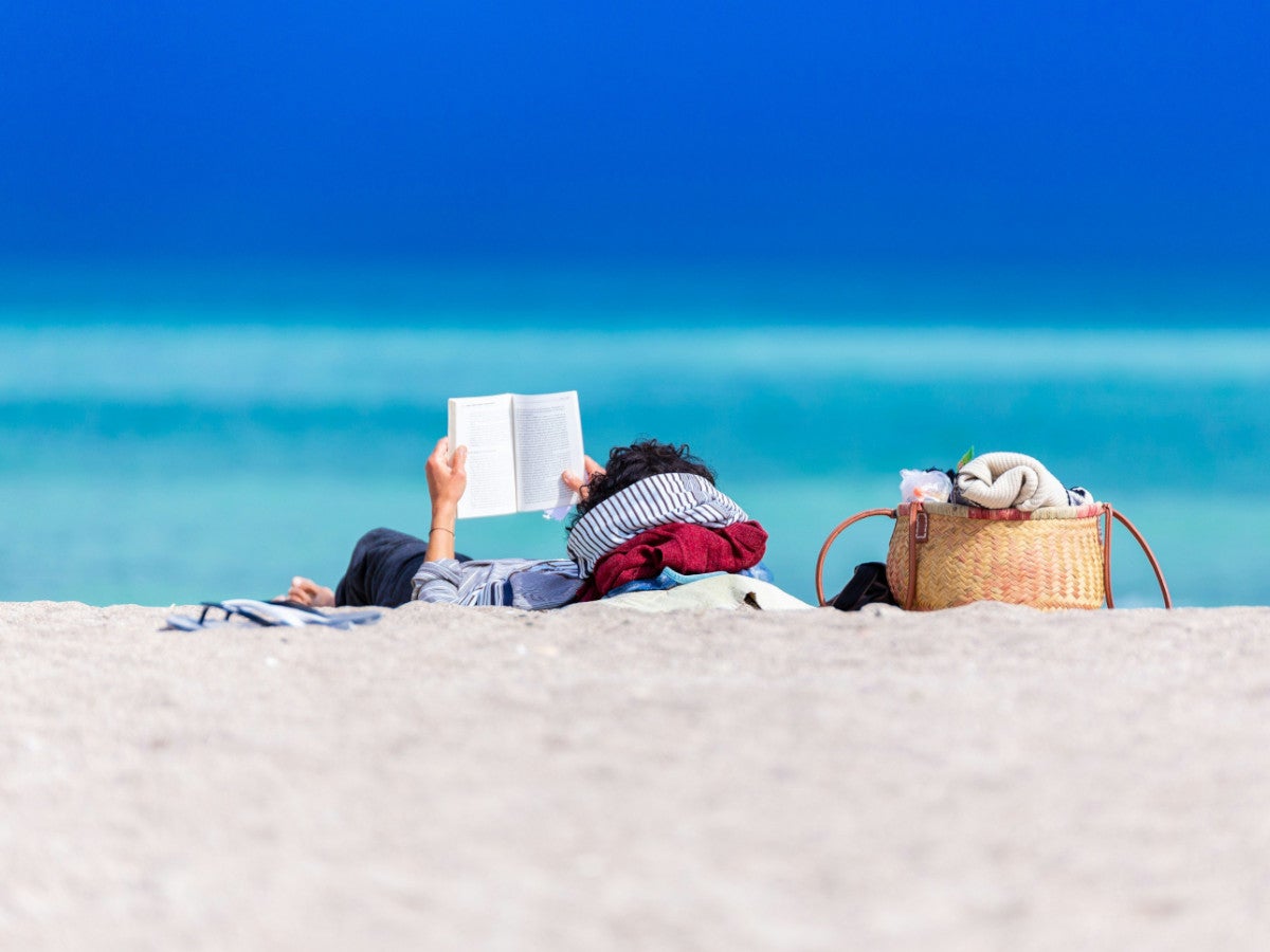 Person liegt mit dem Rücken zur Kamera am Strand und liest ein Buch.